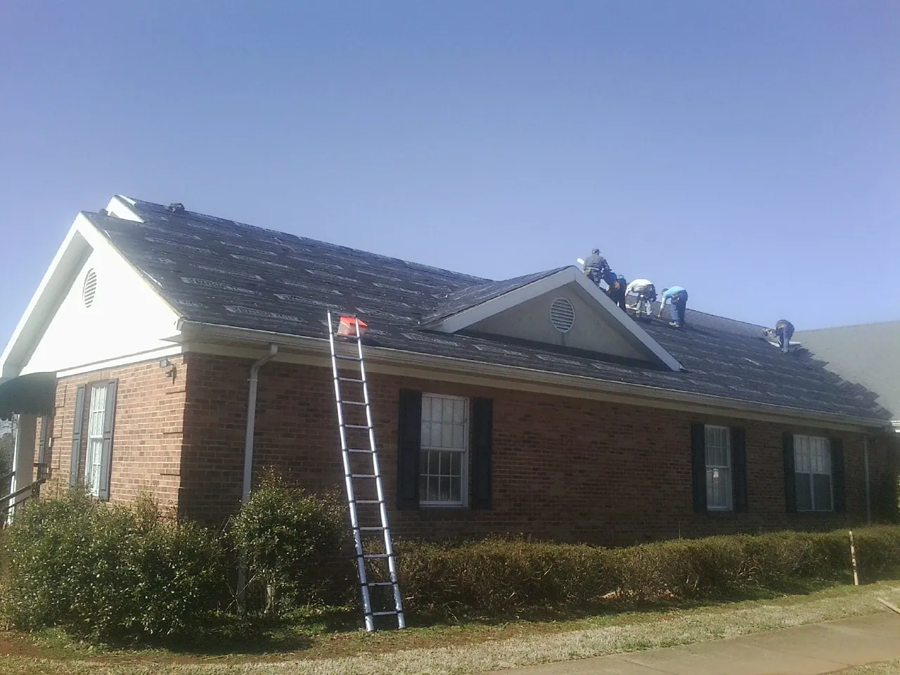Crew tearing shingles off a brick ranch roof with a ladder set — Metal Roof Repair work by our crew in Henrietta