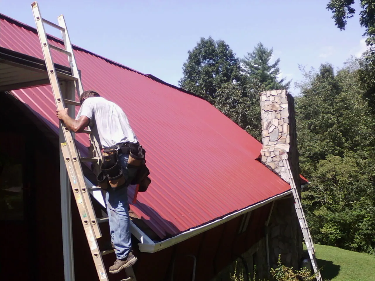 Roofer on a ladder beside a red metal roof and stone chimney — Soffit & Fascia Repair by Roofing Brothers in Henrietta